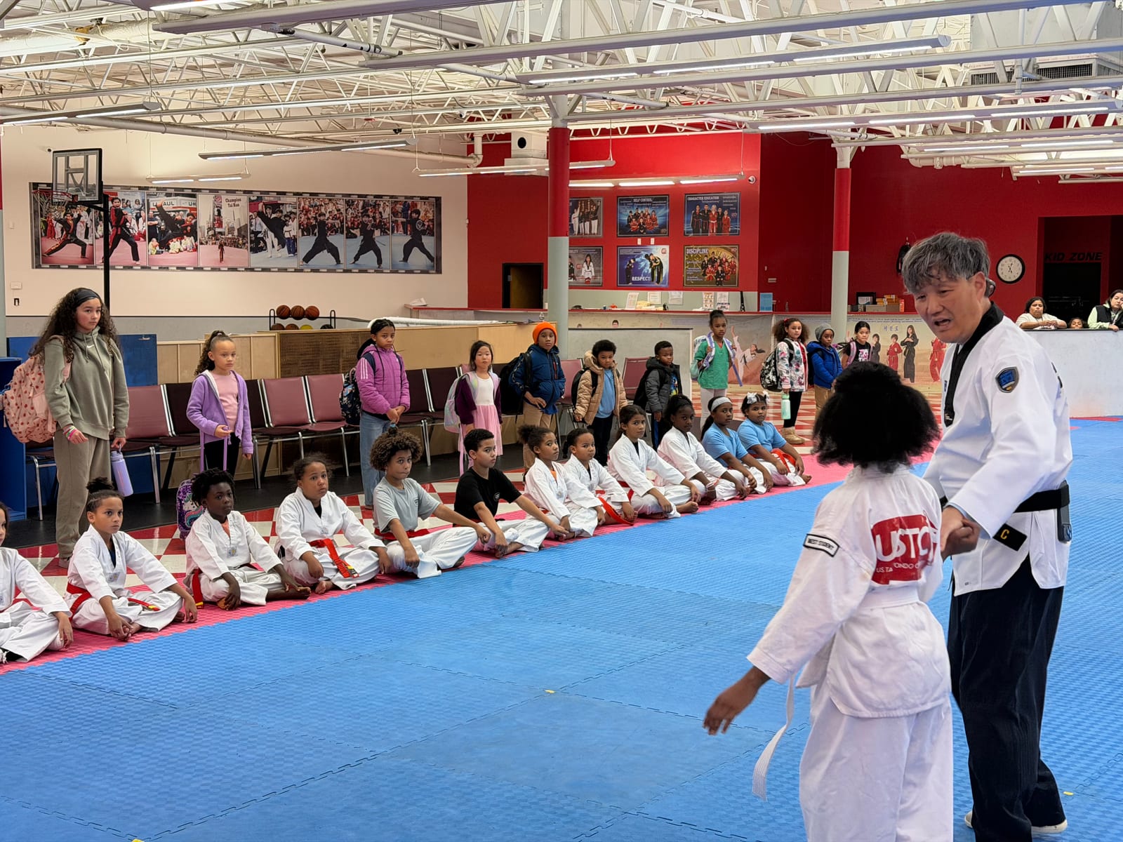Instructor working with a white-belt student on the mats during testing while classmates watch